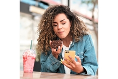 A women sits at a table outside and eats a cookie and drinks a pink icy drink while staring at her phone.